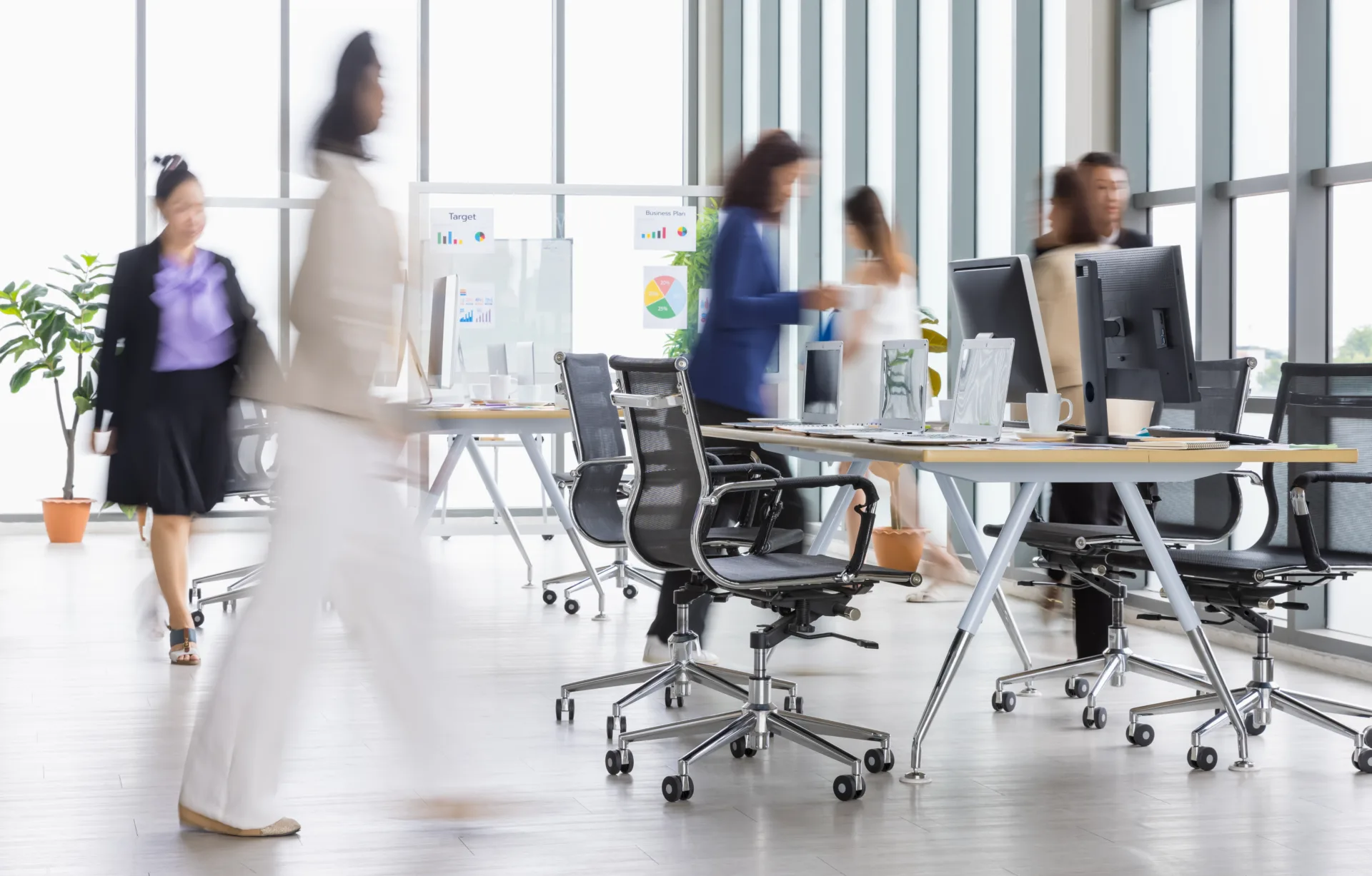 blurry office business women walking in rush in office with multiple glass windows. wooden conference table with laptops and desktop in the center of the office