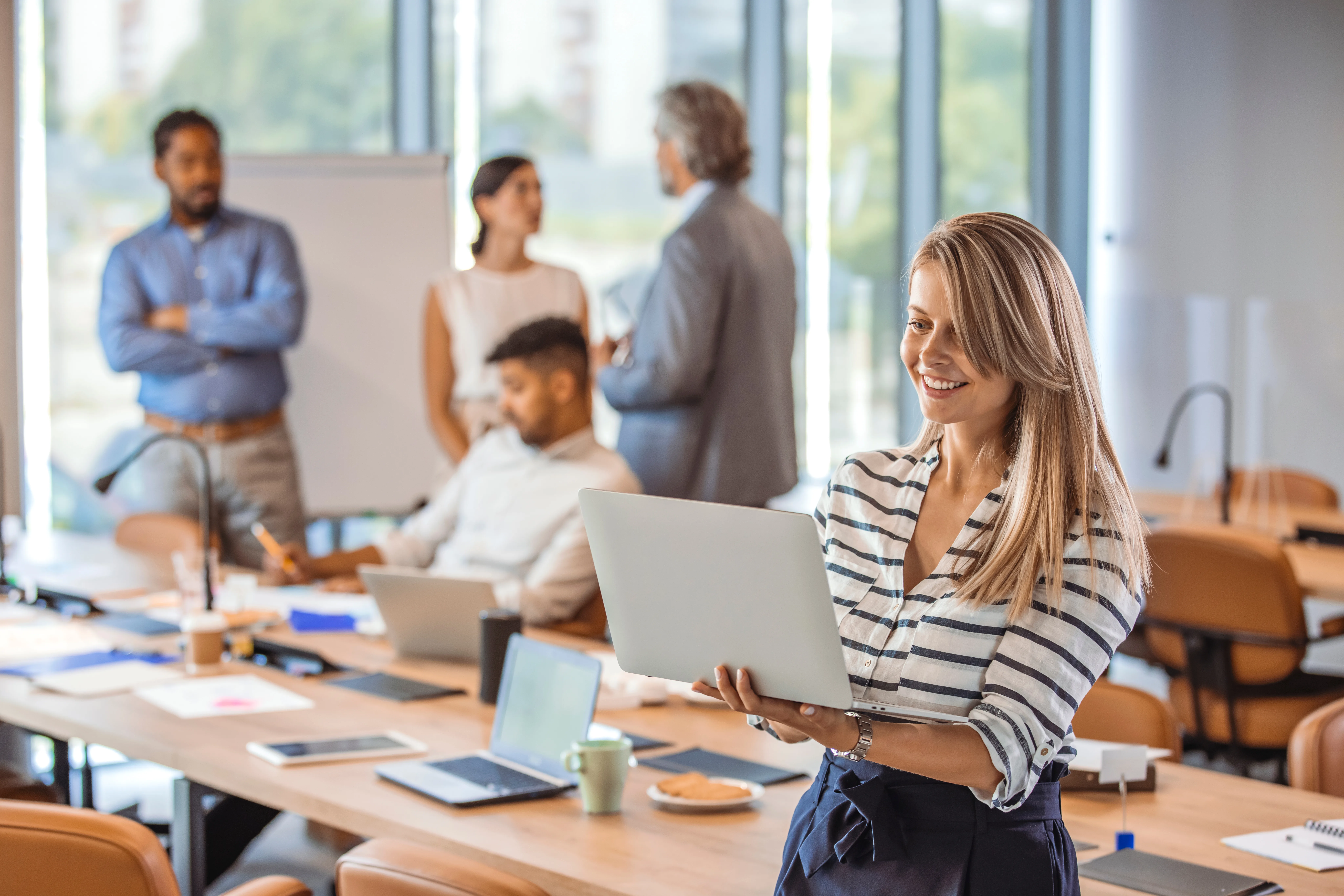 smiling woman with a laptop in office. portrait of a smiling confident young businesswoman holding laptop in hand looking at camera with colleague at background in office