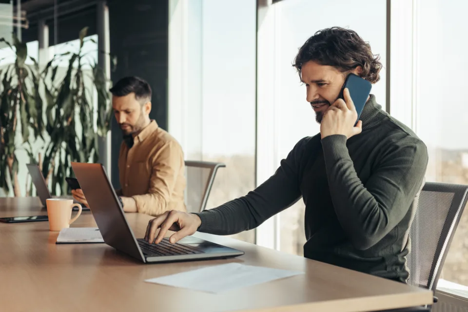 joyful young businessman using laptop, talking on cellphone, working with male colleague at company office, free space