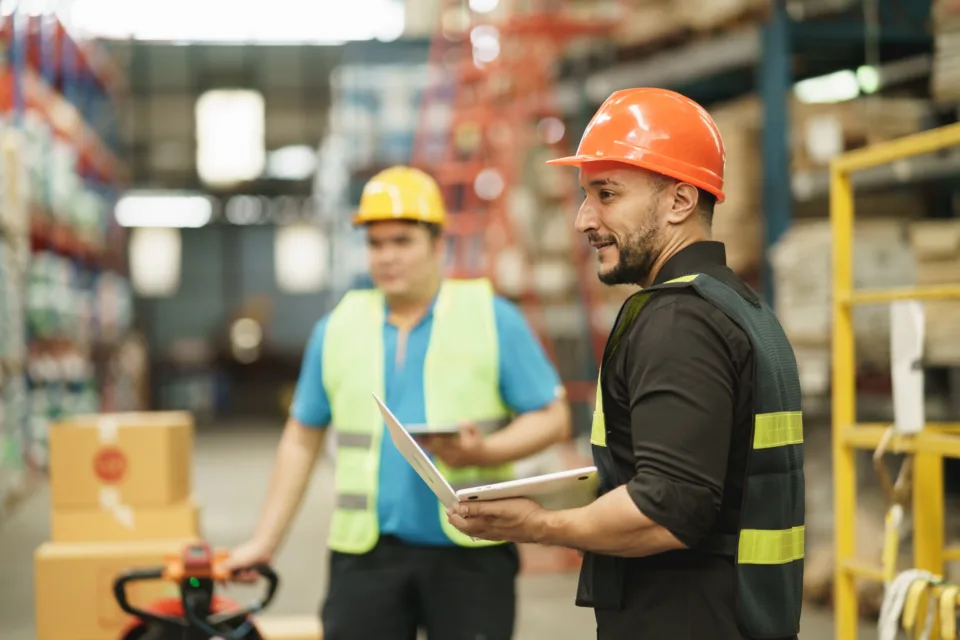 manager man using laptop check stock cardboard on forklift and co worker at warehouse. worker wearing high visibility clothing and hard hat, helmet to checking count up goods or boxes for delivery.